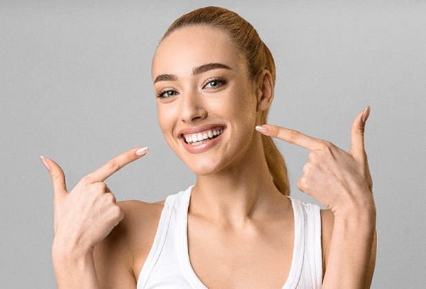A young female wearing a white tank top and pointing to her healthy, beautiful smile after receiving veneers
